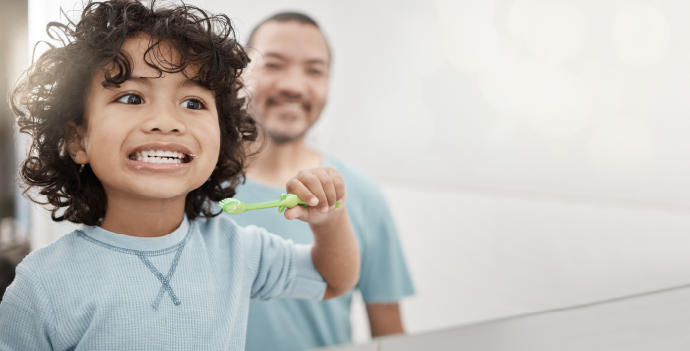 A young child with curly hair brushes their teeth, smiling brightly, while a man, presumably the father, watches with a pleased expression in a brightly lit bathroom.
