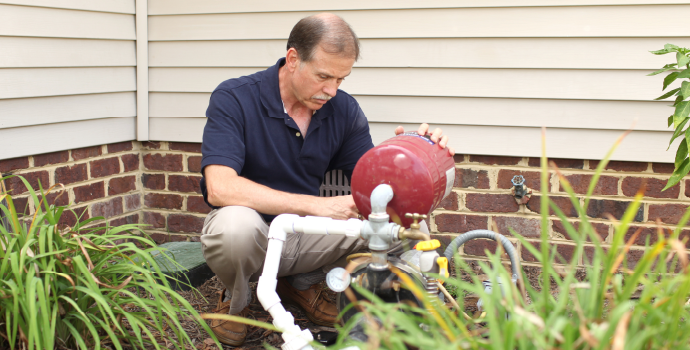 A man kneels while adjusting a red spherical valve on a well water pipe near a brick wall and plants.