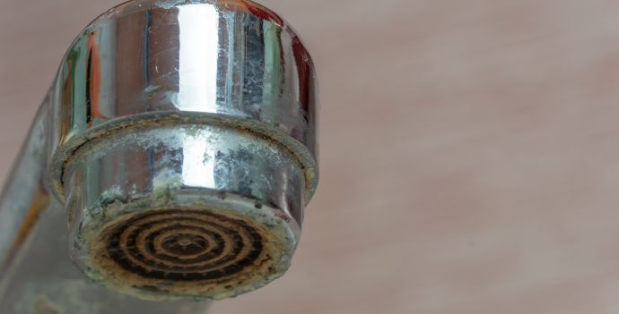 Close-up of a faucet spout with visible limescale buildup on the edges, against a blurred background.