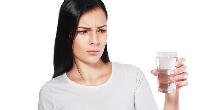 A woman with dark hair is holding a glass of water and looking at it with a concerned expression. She is wearing a white shirt and stands against a neutral background, contemplating the hidden cost of iron in her well water and why removing it might be more affordable than she thinks.