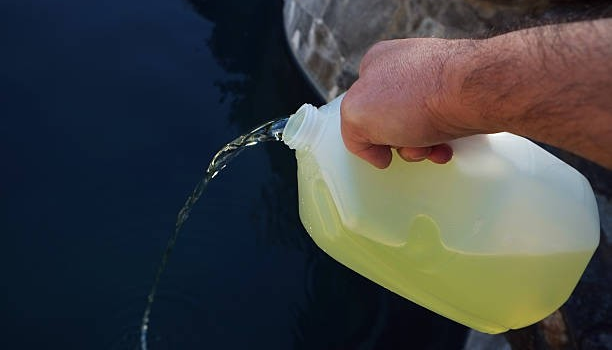 A person is pouring a yellowish liquid from a large plastic jug into a body of water.