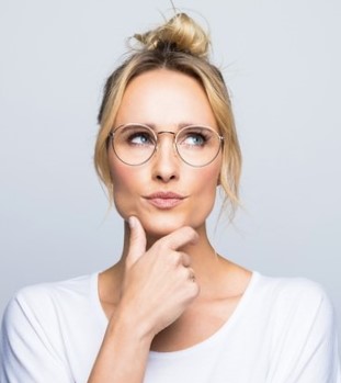 A person with blonde hair in a bun, wearing glasses and a white shirt, looking upwards thoughtfully with one hand on their chin.