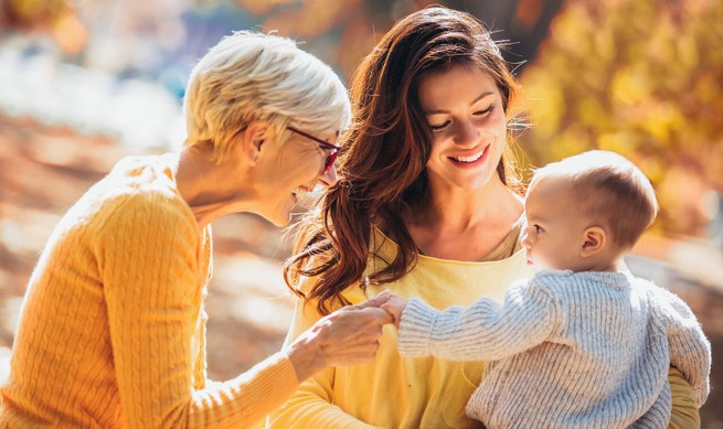 Three generations: an older woman, a younger woman, and a baby are outdoors. The older woman, wearing glasses, and the younger woman, both smile while the baby, in a sweater, engages with them.