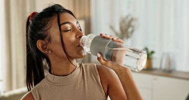 A woman with a ponytail drinks water from a glass bottle in a kitchen with natural light.