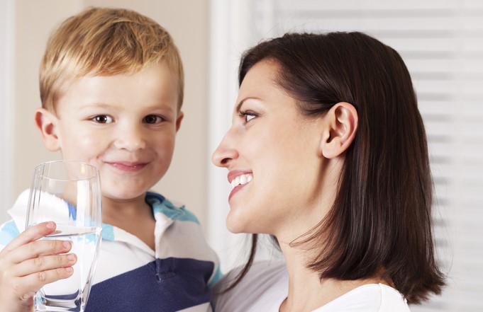 A smiling woman looks at a young child who is holding a glass of water and smiling at the camera.