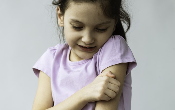 A young girl in a purple shirt touches her upper arm and looks down.