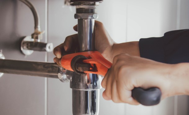 Person using an orange pipe wrench to tighten or loosen a metallic pipe under a sink.