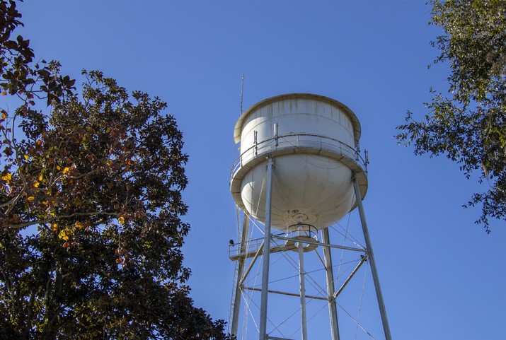 Tall white water tower with metal supports stands against a clear blue sky, framed by trees.