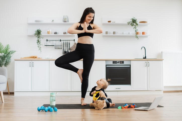 A woman practices tree pose yoga on a mat in a kitchen, while a baby with a toy looks up at her. Nearby are dumbbells, a water bottle, and a laptop.