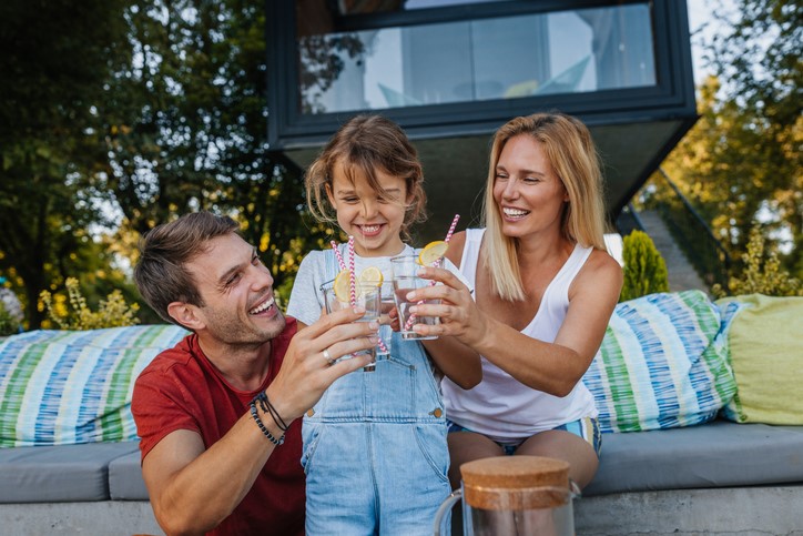 A family of three cheers with drinks on a patio, smiling and sitting on a cushioned bench.