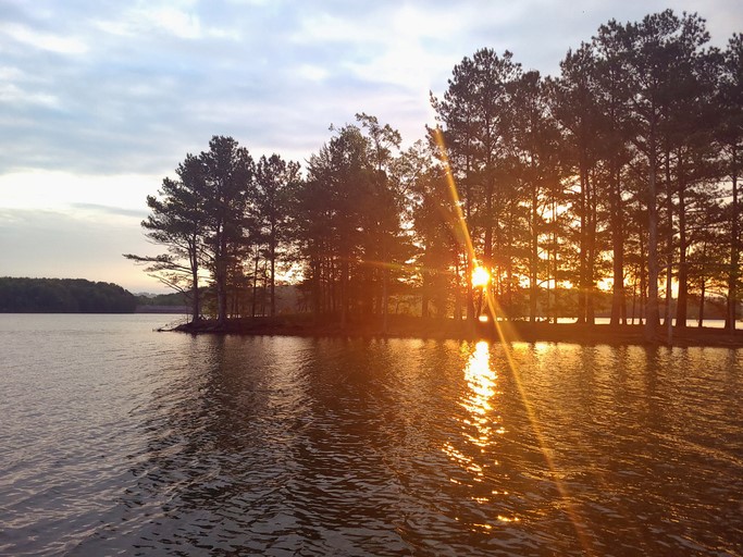 Sunset over a calm lake with a tree-lined shore, reflecting warm light on the water's surface.