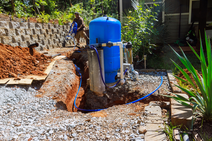 A large blue water tank installed in a garden trench with blue pipes. A person is working nearby, surrounded by gravel, dirt, and green plants.