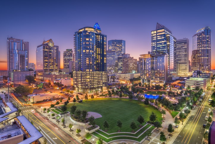 A city skyline at dusk with illuminated skyscrapers surrounding a central green park and busy streets.