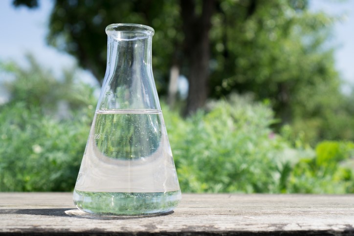A clear glass Erlenmeyer flask filled halfway with transparent liquid sits on a wooden surface outdoors with greenery in the background.