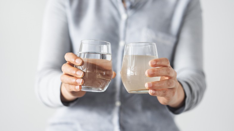 A person holds a clear glass of clean water in one hand and a glass of cloudy, dirty water in the other hand, both at chest height.