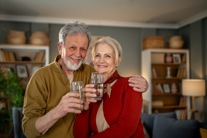 An older man and woman smile while holding glasses of water in a cozy living room, standing close together with bookshelves in the background.