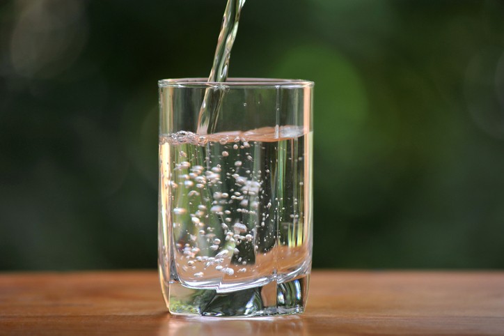 Clear liquid is being poured into a glass on a wooden surface, with bubbles forming and a blurry green background.