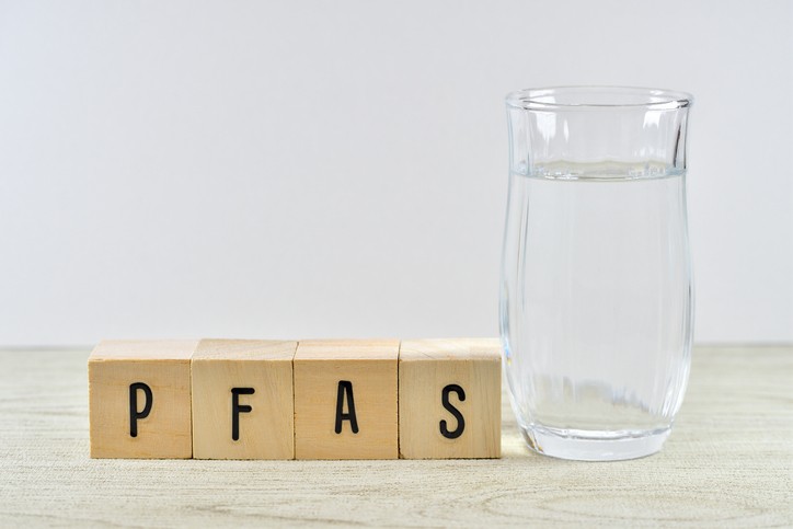 A glass of water stands next to four wooden blocks spelling out "PFAS" on a light-colored surface.