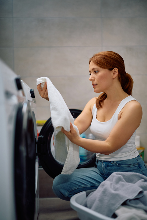 A woman kneels by a washing machine, holding a white towel and inspecting it, with a laundry basket of clothes in front of her.