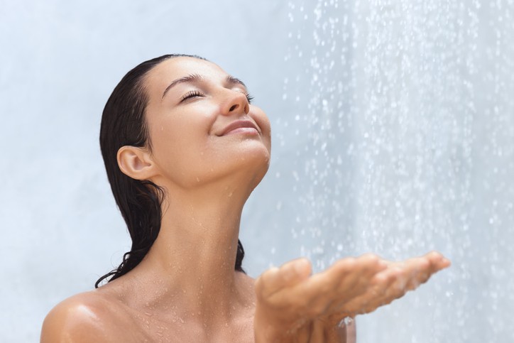 A woman with wet hair stands under a shower, eyes closed and smiling, with one hand raised to catch falling water droplets.
