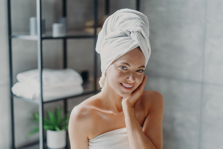 Woman wrapped in a white towel with a towel on her head stands in a modern bathroom, smiling and touching her face. Shelves and a potted plant are in the background.