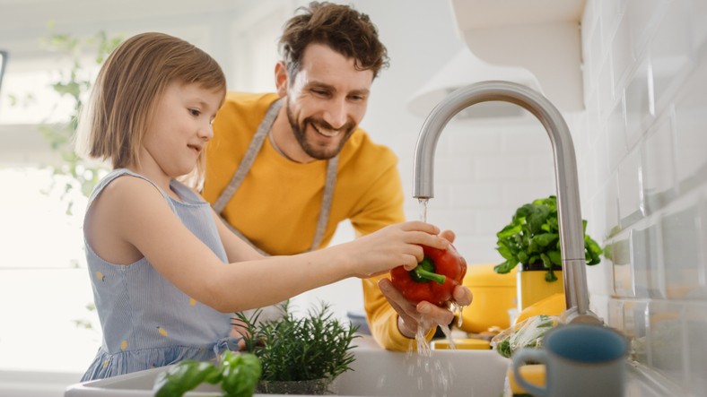 An adult and a child wash a red bell pepper under a kitchen faucet surrounded by fresh vegetables and herbs.