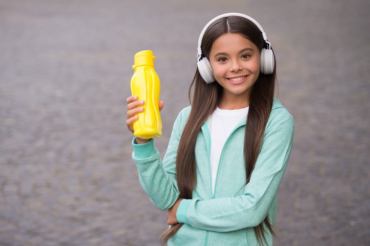 A smiling girl wearing headphones and a light green jacket holds up a yellow water bottle while standing outdoors.