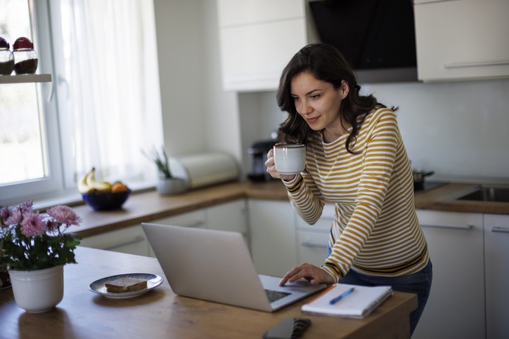 A woman in a striped shirt stands in a kitchen, holding a mug and looking at a laptop. A notebook, pen, and breakfast plate are on the counter.