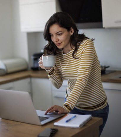A woman in a striped shirt stands in a kitchen, holding a mug and looking at a laptop, with a notepad and pen on the counter.