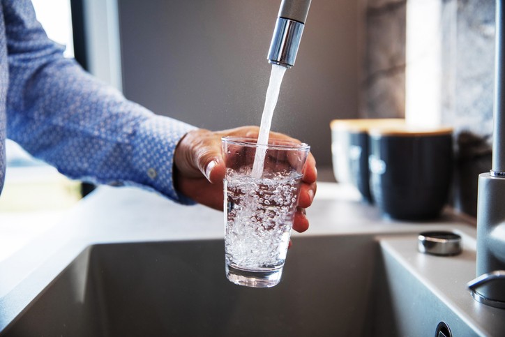 A person fills a clear glass with water from a kitchen faucet over a sink.