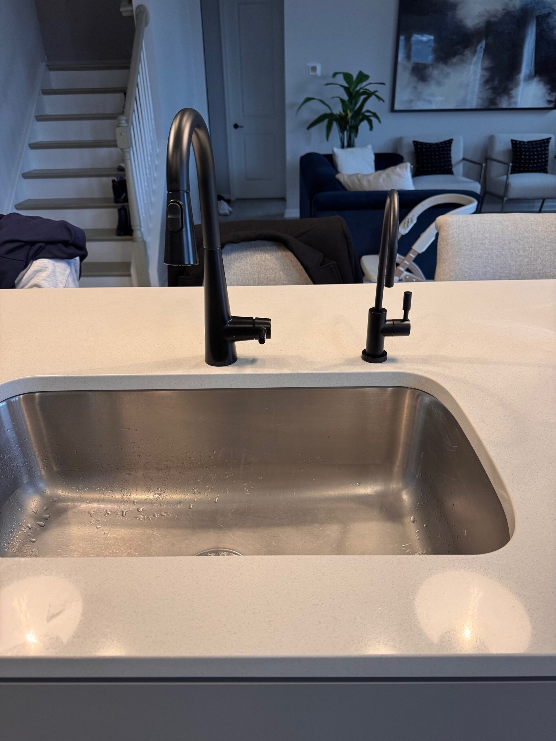 A modern stainless steel kitchen sink with two matte black faucets on a white countertop; a staircase, living area, and plant are visible in the background.