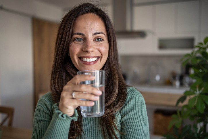 A woman with long brown hair, wearing a green sweater, smiles while holding a glass of water in a modern kitchen.