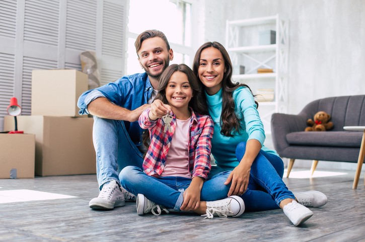 A smiling man, woman, and girl sit on the floor of a living room with moving boxes; the girl holds up a set of keys.