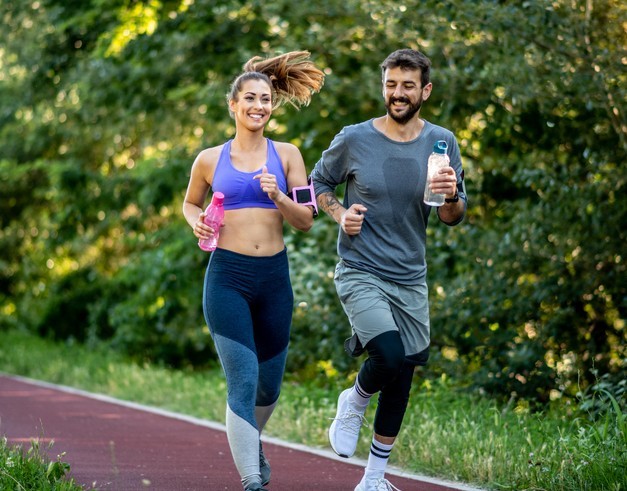 A woman and a man jogging together on an outdoor track, holding water bottles, with trees in the background.