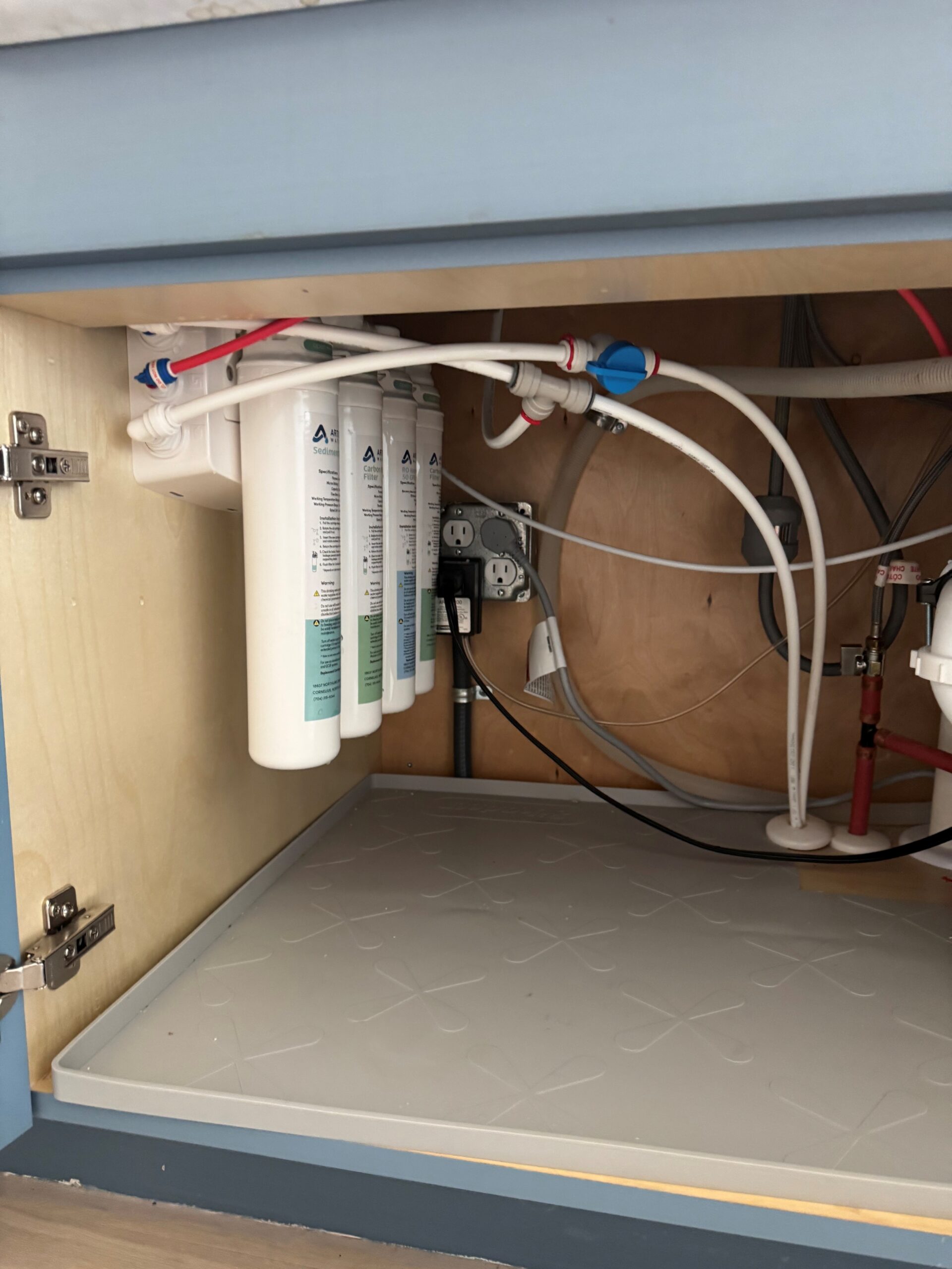 Three water filter cartridges and various tubes are mounted under a sink, showcasing a Water Filtration Mooresville NC setup with electrical outlets and plumbing visible against the cabinet wall.