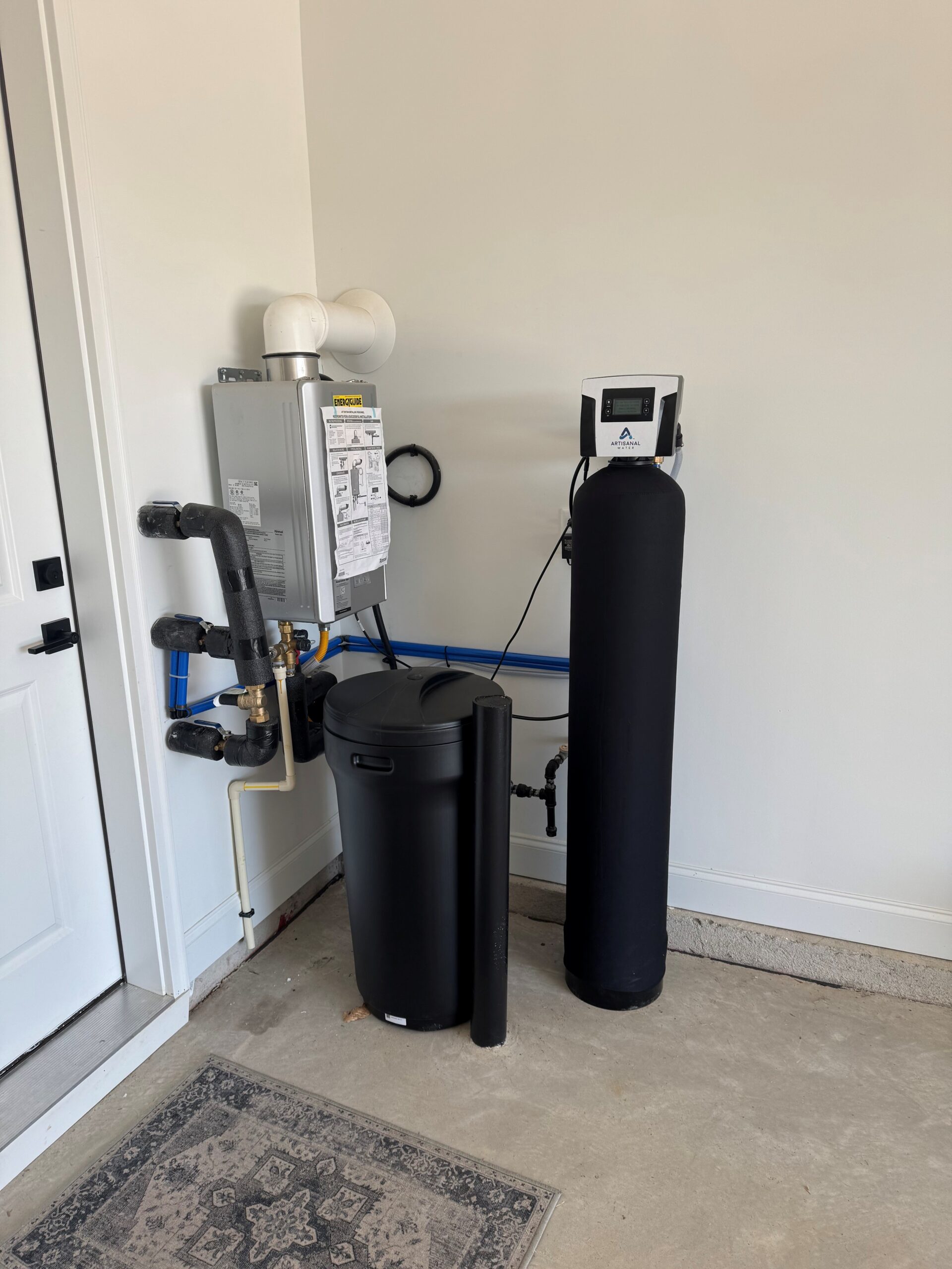 Well Water Treatment System with a water softener and filtration system installed in a garage corner, featuring pipes connected to a heater unit and a control panel on top of the tall black tank.