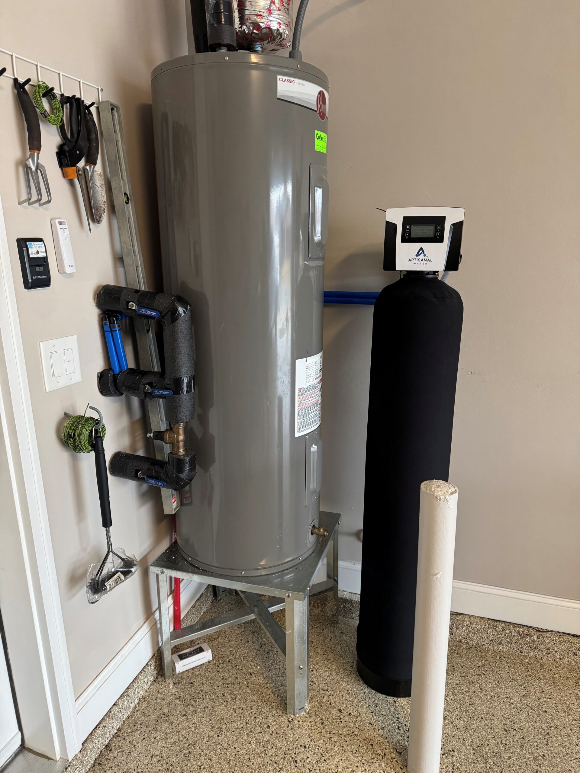 A gray water heater on a metal stand next to a black water filtration tank in a room with tools hanging on the wall.