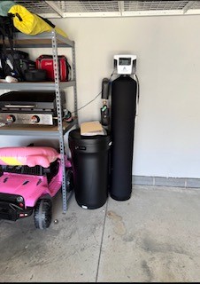 A storage shelf with a pink toy jeep parked below it, next to a black water softener tank and a black brine tank.